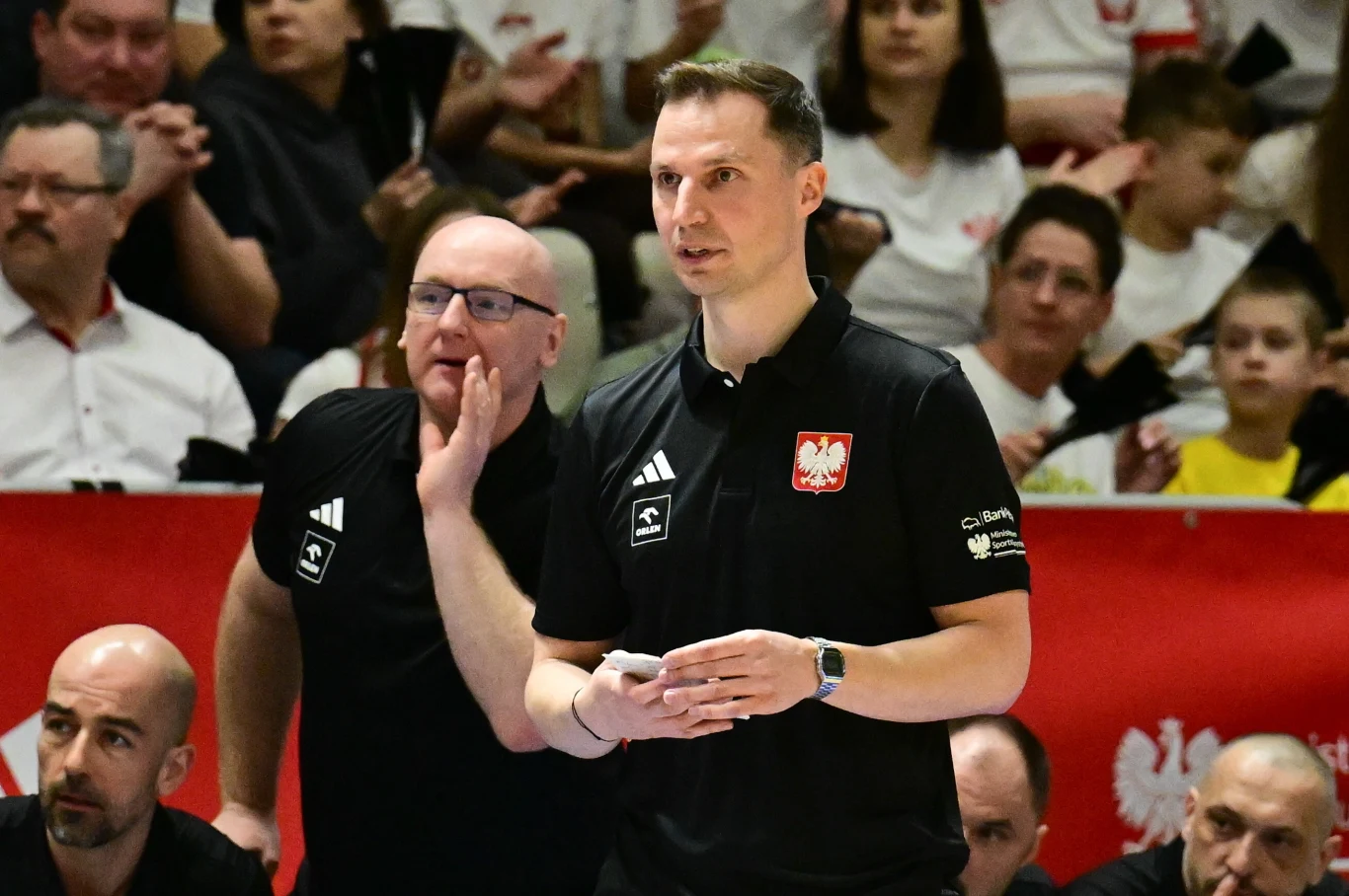 Dwóch trenerów w czarnych koszulkach z polskim godłem na piersi stoi na tle kibiców podczas meczu siatkarskiego; jeden z nich trzyma notatki, drugi gestykuluje, wokół nich widoczni są inni członkowie sztabu i skupiona publiczność.