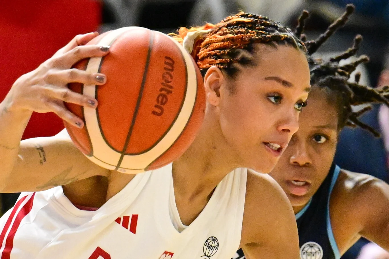 Koszykarka w białym stroju trzyma piłkę do koszykówki blisko głowy w trakcie dynamicznej akcji, za jej plecami widoczna jest zawodniczka drużyny przeciwnej w ciemnoniebieskim stroju.