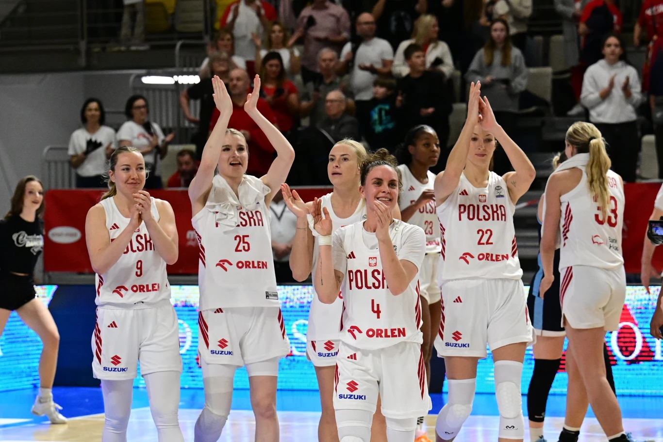 Koszykarki reprezentacji Polski w białych strojach z napisem 'Polska' i numerami na froncie, stoją razem na boisku, klaszcząc i uśmiechając się, w tle widać kibiców na trybunach.