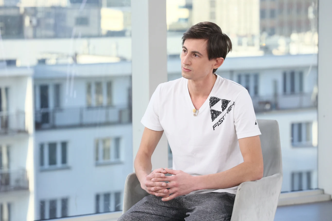 Młody mężczyzna o ciemnych włosach siedzi na jasnym krześle w nowoczesnym wnętrzu z dużymi oknami, ubrany w białą koszulkę z czarnym nadrukiem i szare spodnie, z dłońmi splecionymi na kolanach, patrzy w bok z zamyślonym wyrazem twarzy.