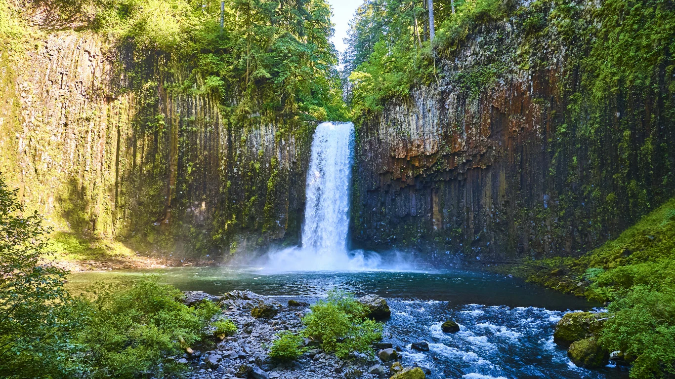 Wysoki wodospad spływający pionowo ze skalnej ściany otoczonej bujną zielenią, woda wpada do spokojnego, niebieskiego jeziora otoczonego skałami i gęstymi krzewami.