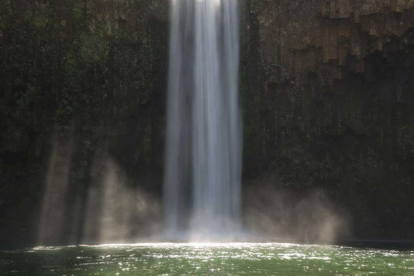 Wysoki wodospad spadający pionowo do zielonkawej wody, otoczony ciemnymi, skalistymi ścianami z lekką mgiełką unoszącą się u podstawy kaskady.