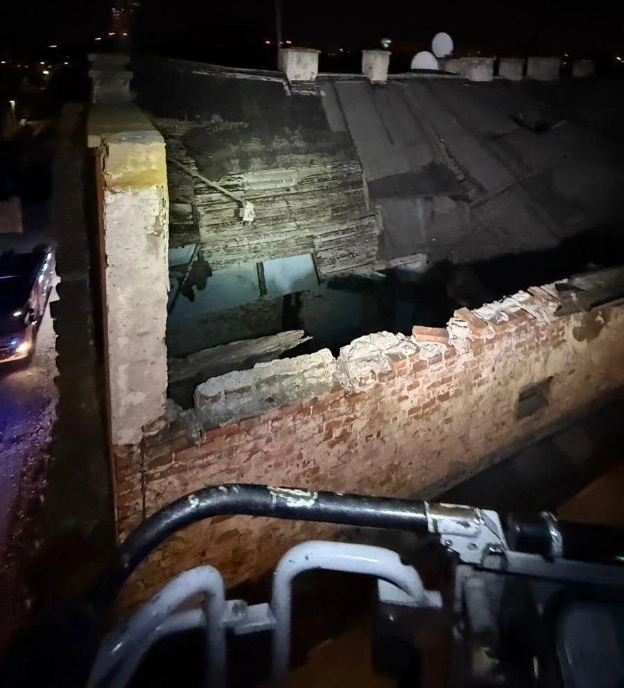 Zawalony fragment dachu i ściany starego, ceglanego budynku oświetlony w nocy, widoczne uszkodzenia konstrukcji oraz ruiny wewnętrzne, na pierwszym planie metalowy fragment pojazdu lub podnośnika strażackiego.