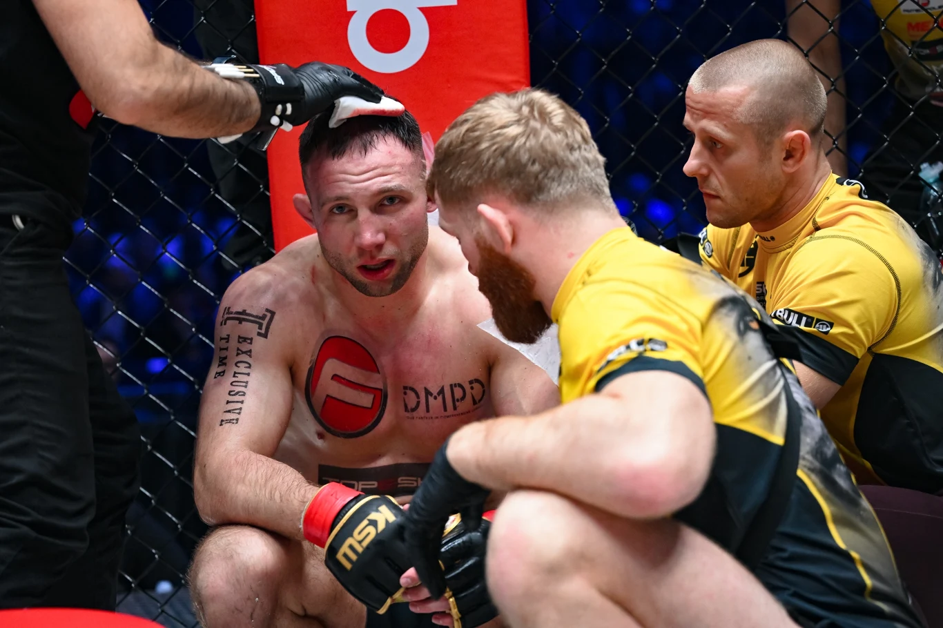 Zawodnik MMA siedzi w narożniku oktagonu z widocznymi śladami zmęczenia na twarzy, otoczony przez członków swojego zespołu udzielających mu wskazówek i wsparcia w trakcie przerwy między rundami.