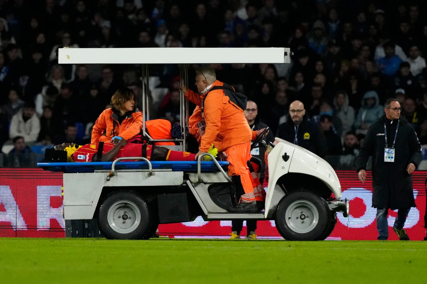 Lameck Banda znoszony z boiska Ratownicy medyczni w pomarańczowych kombinezonach transportują osobę na noszach na specjalnym wózku medycznym po murawie stadionu, w tle widoczna grupa widzów oraz personelu technicznego.