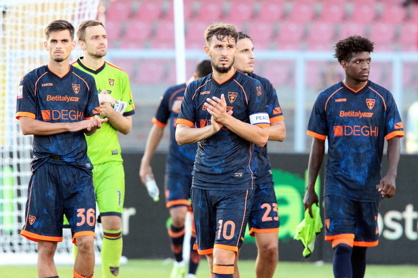 Piłkarze Lecce, wśród nich Filip Marchwiński (z numerem 36) Pięciu piłkarzy w ciemnych strojach drużyny piłkarskiej z numerami na spodenkach wychodzi z murawy, jeden z nich klaszcze, a pozostali wyglądają na zamyślonych lub skupionych; w tle widoczne trybuny stadionu.