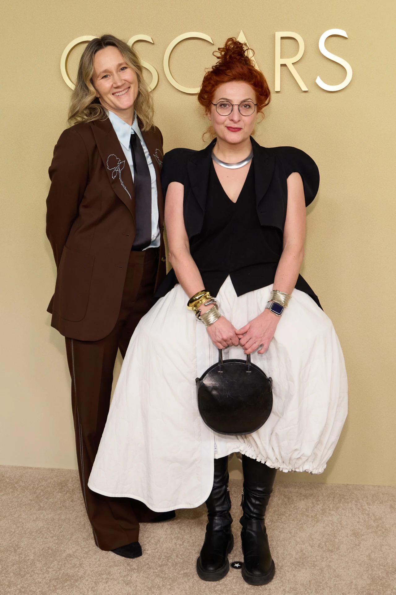 Dwie kobiety pozujące na tle napisu 'OSCAR', jedna ubrana w brązowy garnitur z niebieską koszulą, druga w oryginalnej stylizacji z białą, szeroką spódnicą, czarną górą, licznymi bransoletkami oraz okrągłą torebką; obie stoją blisko siebie uśmiechnięte.