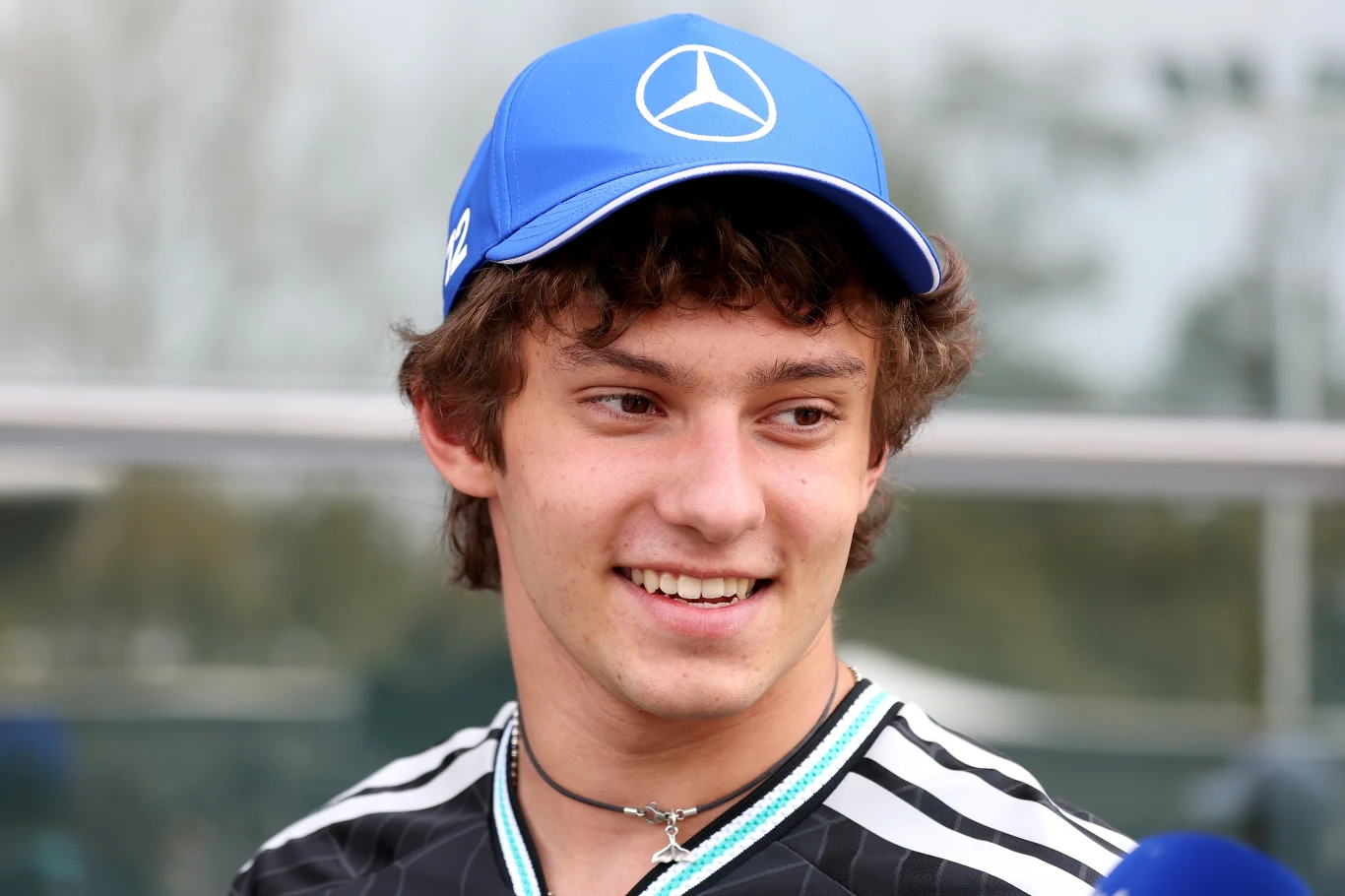 Młody mężczyzna w niebieskiej czapce z logo Mercedesa, uśmiechnięty, ubrany w sportową koszulkę, stoi na zewnątrz przed przeszklonym budynkiem.