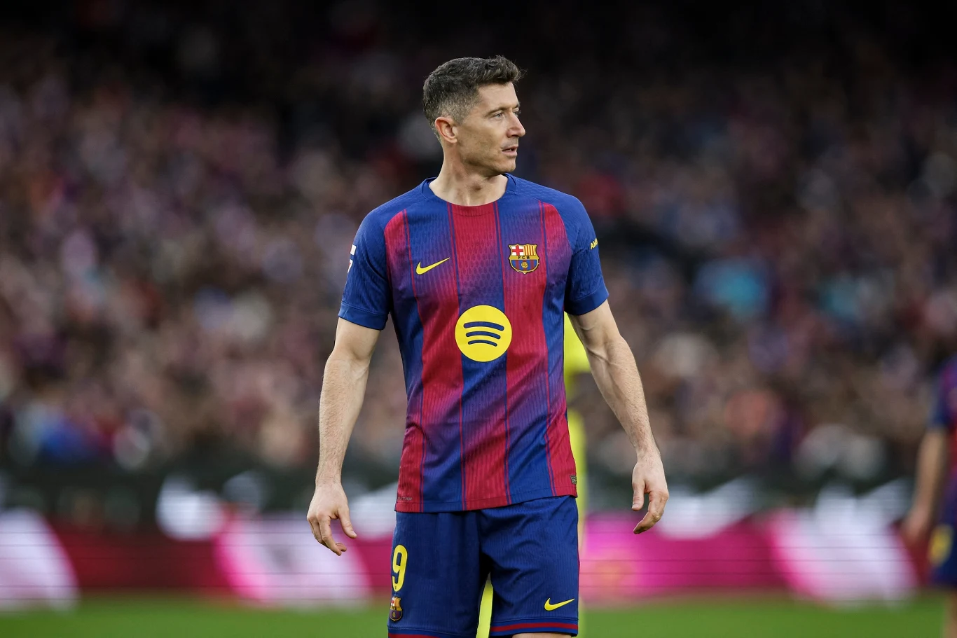 Piłkarz w koszulce FC Barcelony z numerem 9 pośrodku boiska podczas meczu, na tle rozmytego tłumu kibiców na trybunach.