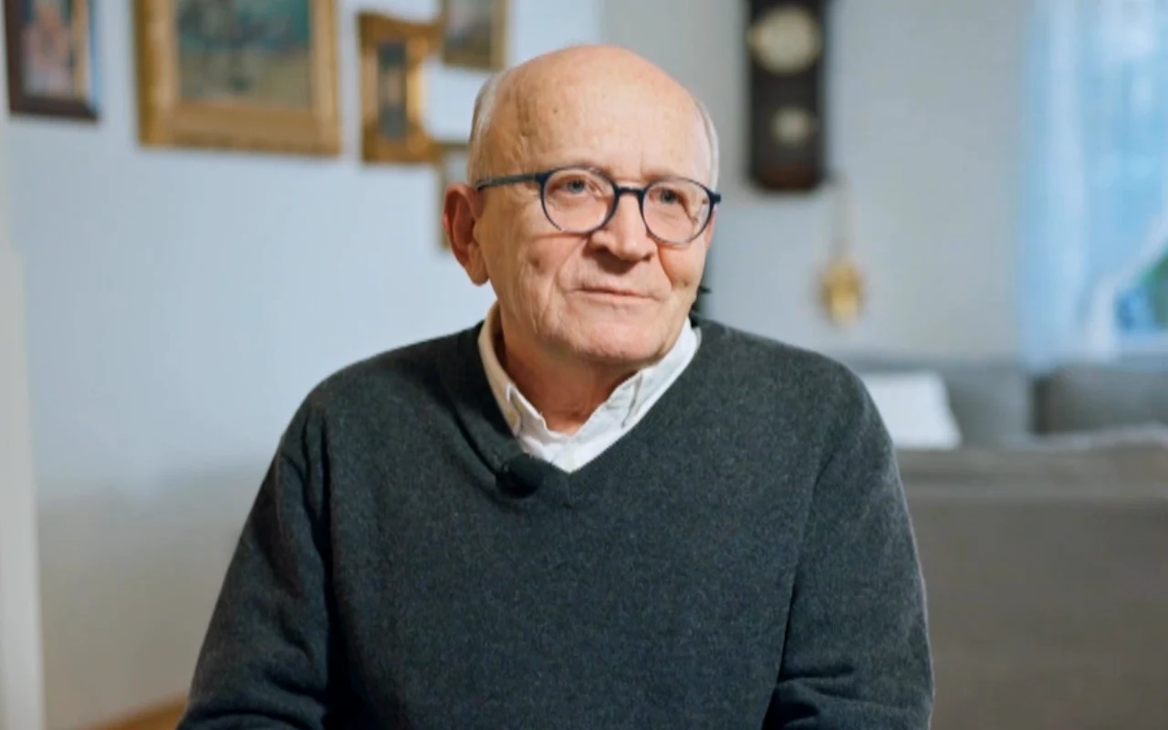 Starszy mężczyzna w okularach i ciemnoszarym swetrze siedzi w jasnym, przytulnym pomieszczeniu; na ścianie za nim widoczne są obrazy i zegar ścienny.
