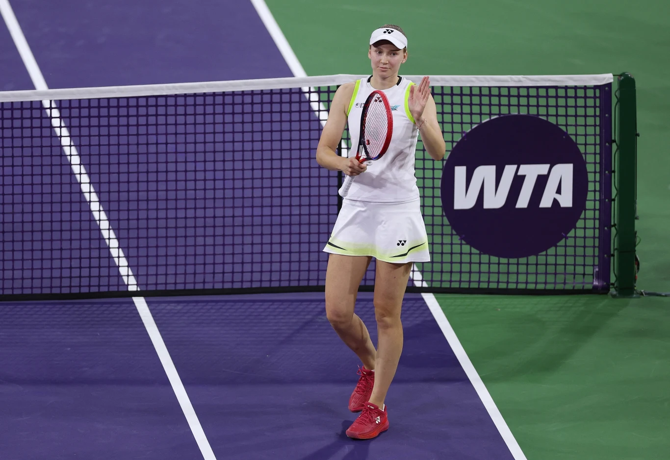 Tenisistka w białym stroju i czerwonych butach stoi na korcie przed siatką, trzyma rakietę i unosi rękę w geście pozdrowienia; w tle logo WTA na siatce.