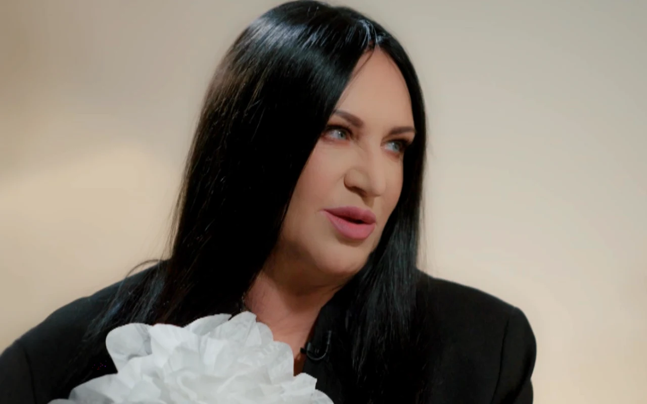 Kobieta o długich, czarnych włosach ubrana w czarną marynarkę z dużą, białą ozdobą przypominającą kwiat, patrząca w bok na jasnym tle.