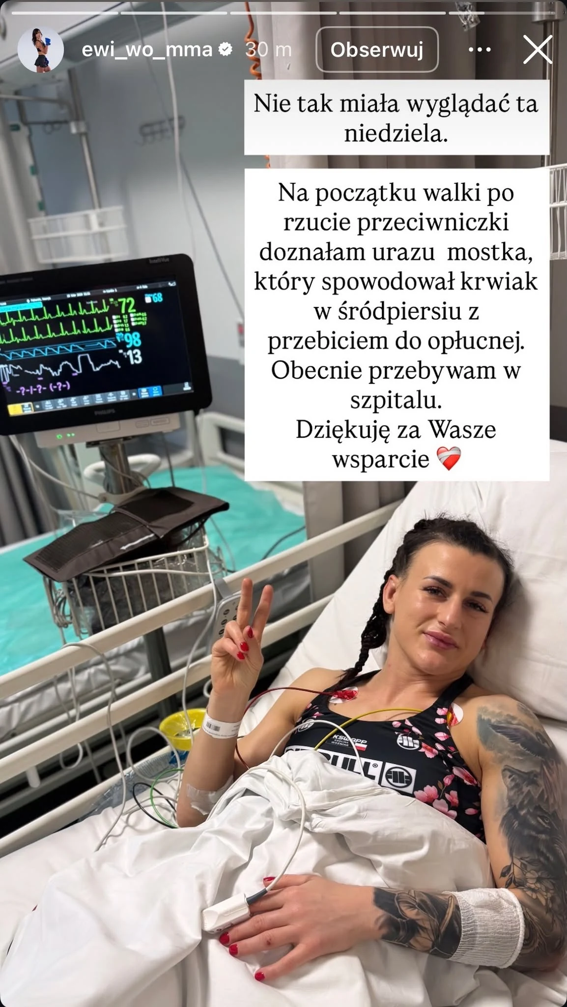 Kobieta leży na szpitalnym łóżku z opaską na ramieniu, pokazując znak pokoju, obok znajduje się monitor medyczny z widocznymi parametrami życiowymi, w tle sprzęt szpitalny i koszyk na kółkach; na zdjęciu umieszczony jest tekst informujący o urazie dozn...