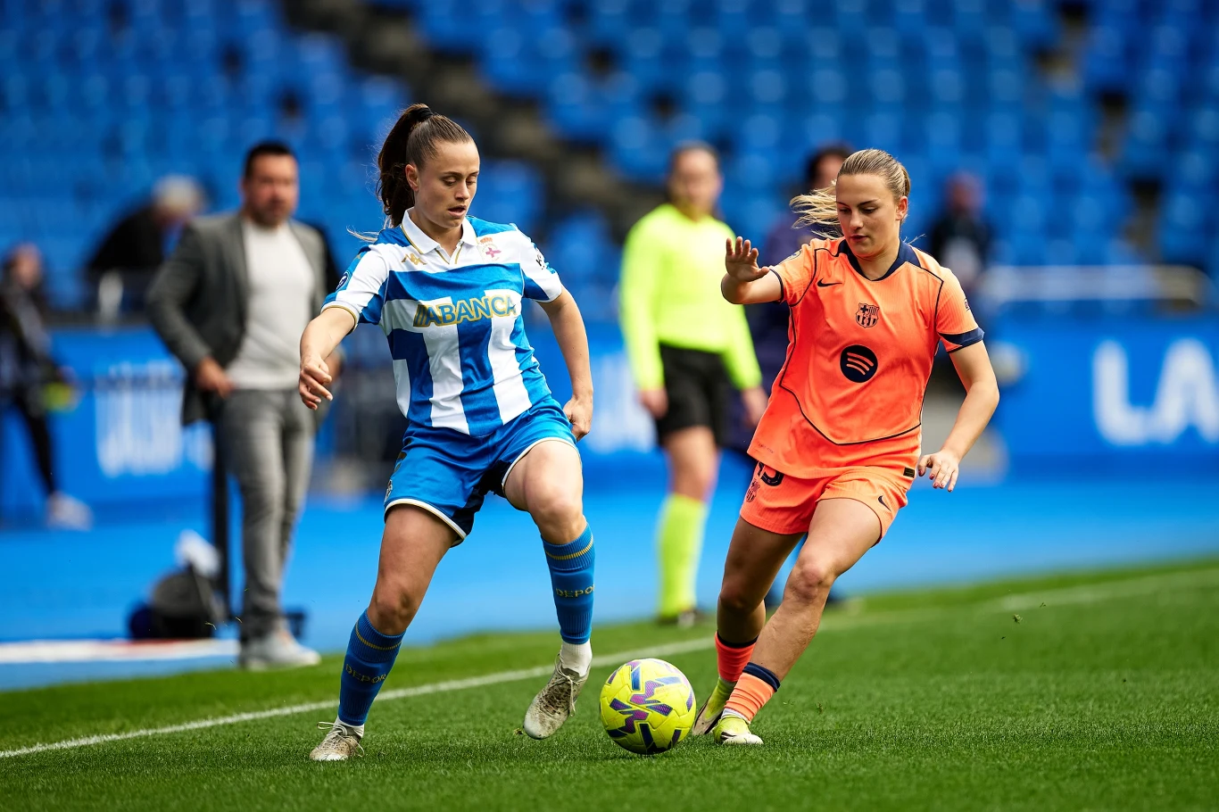 Dwie piłkarki w trakcie rywalizacji o piłkę na murawie stadionu, jedna w niebiesko-białym stroju, druga w pomarańczowym, w tle rozmyta publiczność i sędzia w żółtym stroju.