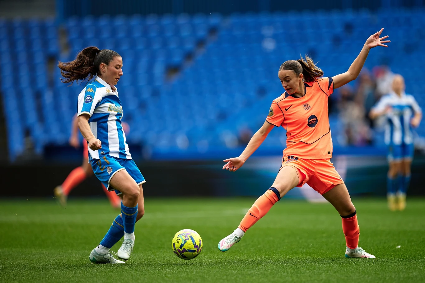 Dwie piłkarki rywalizują o piłkę na murawie; jedna ubrana w biało-niebieski strój, druga w pomarańczowy, obie skoncentrowane na grze na tle pustych niebieskich trybun stadionu.