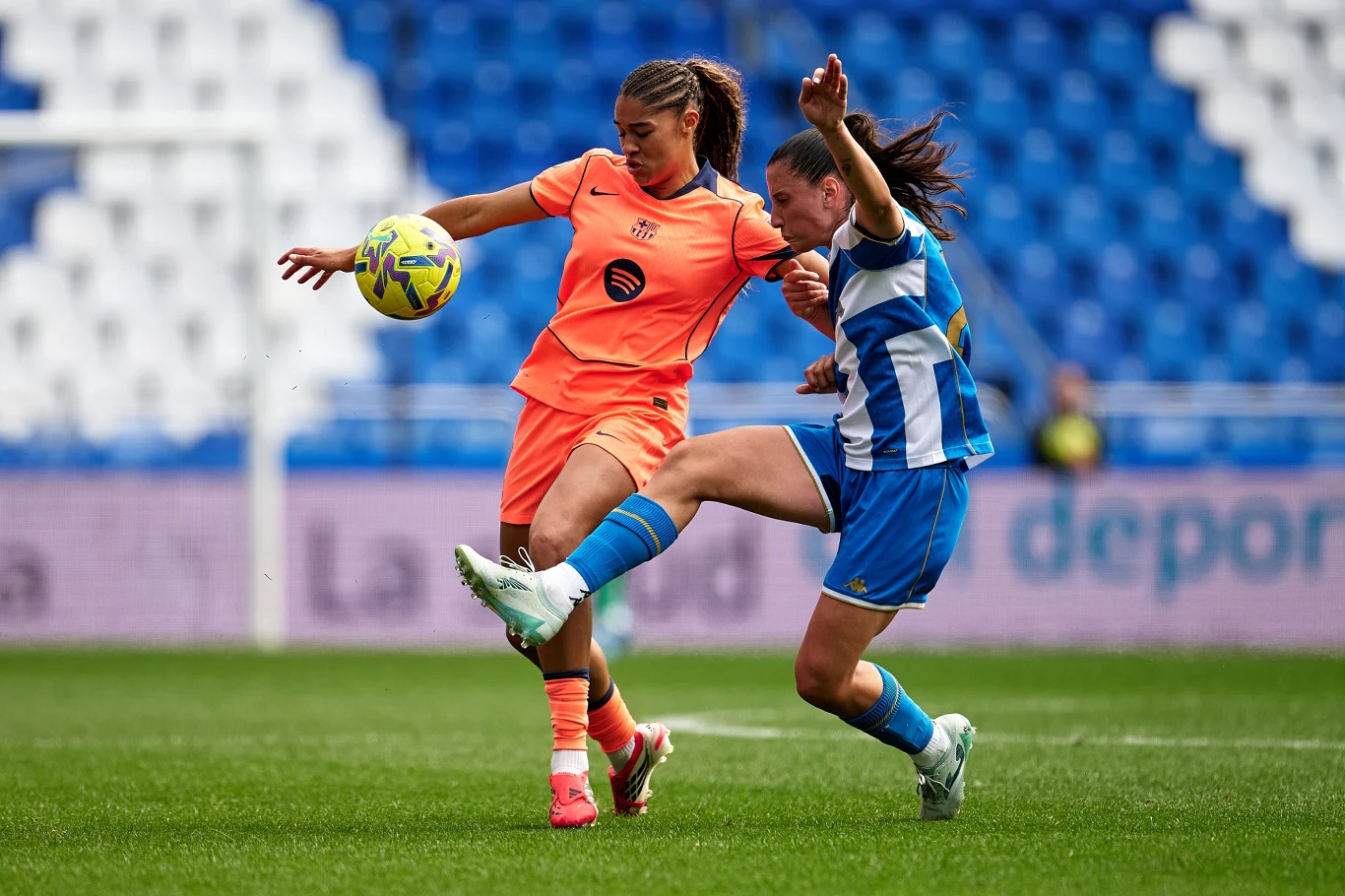 Dwie piłkarki rywalizują o piłkę na boisku, jedna w pomarańczowym stroju wykopuje piłkę, druga w niebiesko-białym stroju próbuje ją zablokować energicznym ruchem nogi.
