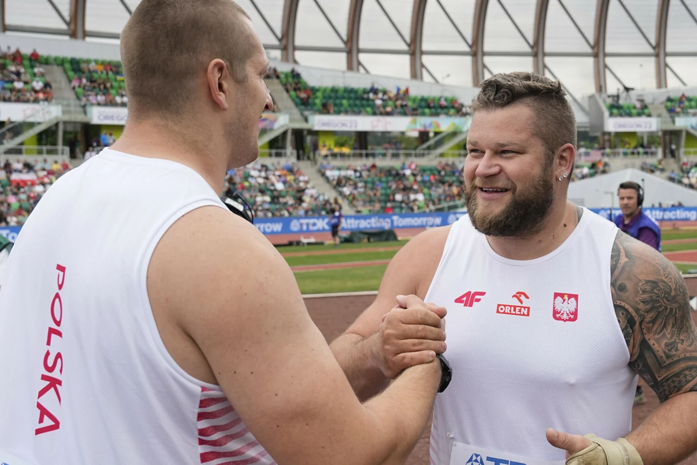 Dwóch lekkoatletów w biało-czerwonych strojach z napisem 'Polska' mocno ściska sobie dłonie na stadionie lekkoatletycznym, wymieniając uśmiechy i gratulacje po udanym występie sportowym.