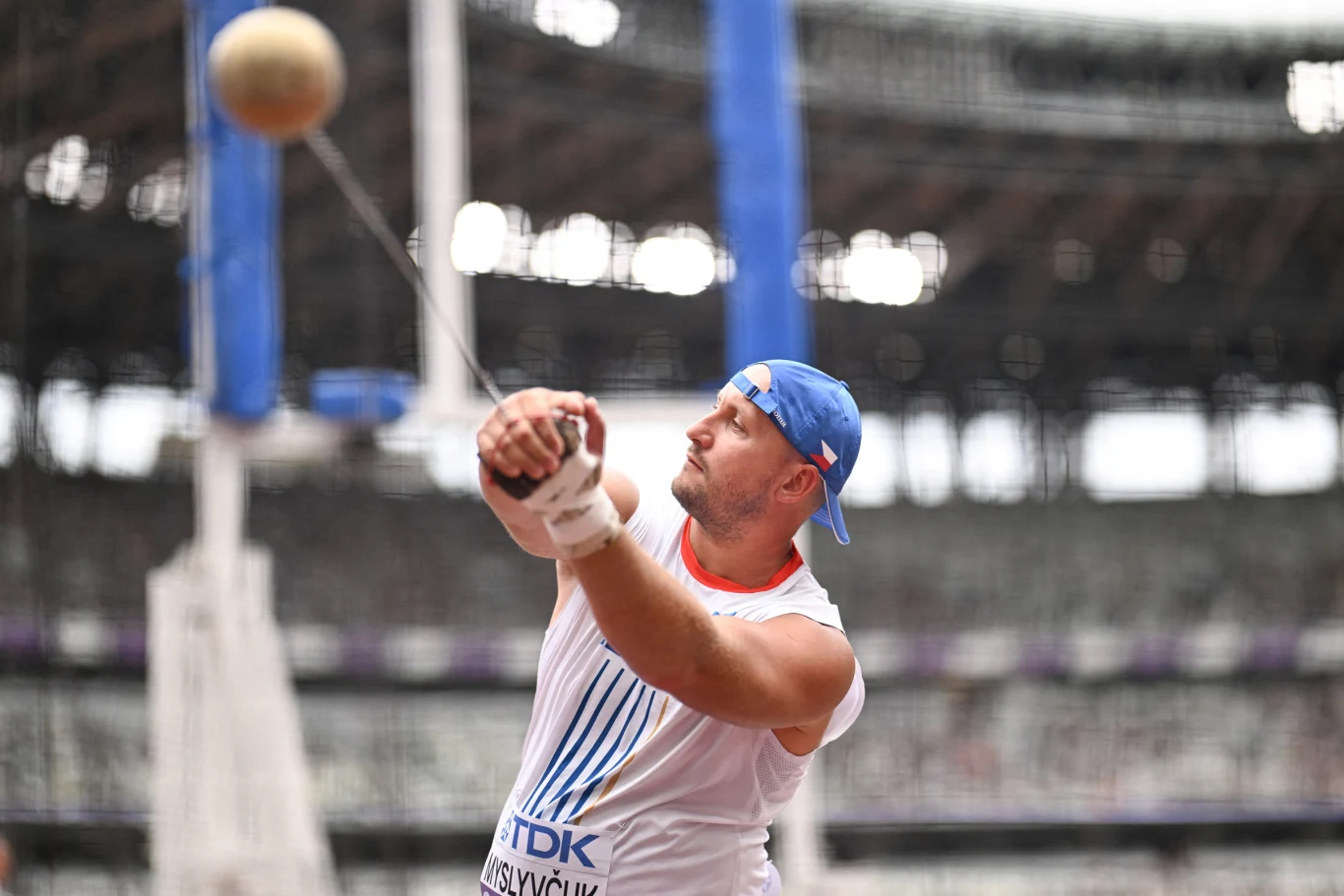 Lekkoatleta w białej koszulce i niebieskiej czapce wykonuje rzut młotem na stadionie, koncentrując się na technice podczas dynamicznego ruchu obrotowego.