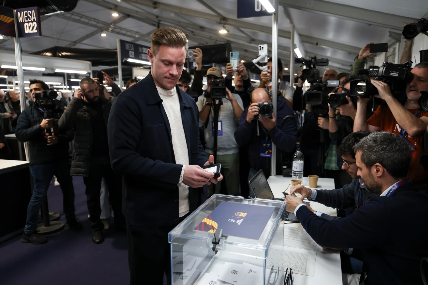 Mężczyzna wrzuca głos do przezroczystej urny wyborczej, przy której siedzi dwóch członków komisji z dokumentami, wokół zgromadzeni liczni fotoreporterzy i kamery rejestrujące wydarzenie.