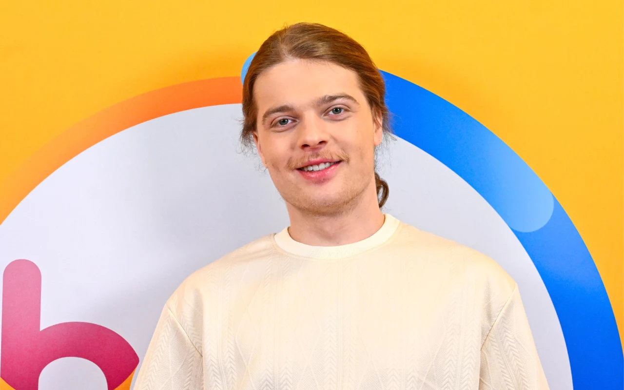 Młody mężczyzna z jasnymi włosami spiętymi w kitkę, uśmiecha się subtelnie, ubrany w kremową bluzę, stoi na tle żółtej ściany z kolorowym, okrągłym logo w odcieniach niebieskiego i czerwonego.