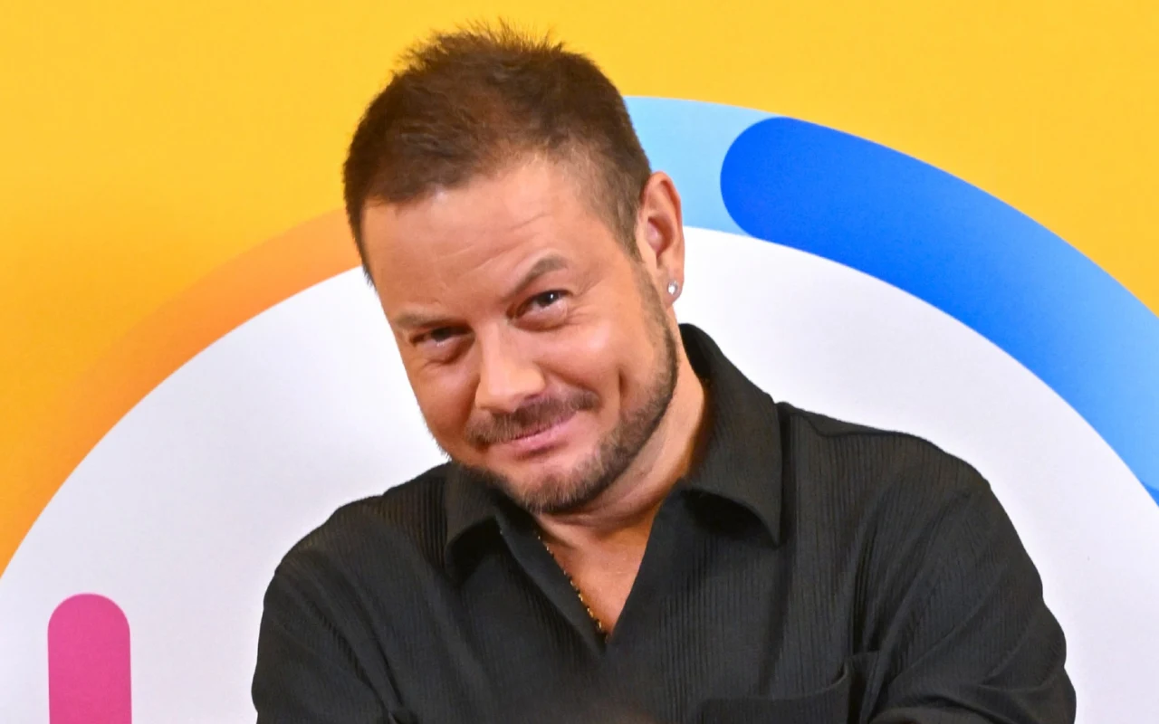 Bartek Wrona w "halo tu polsat" Mężczyzna o krótkich, ciemnych włosach ubrany w czarną koszulę z lekko rozpiętym kołnierzykiem stoi na tle kolorowej grafiki w kształcie okręgu z elementami niebieskimi, pomarańczowymi i różowymi, lekko uśmiechnięty, z widocznym kolczykiem w lewym uchu.