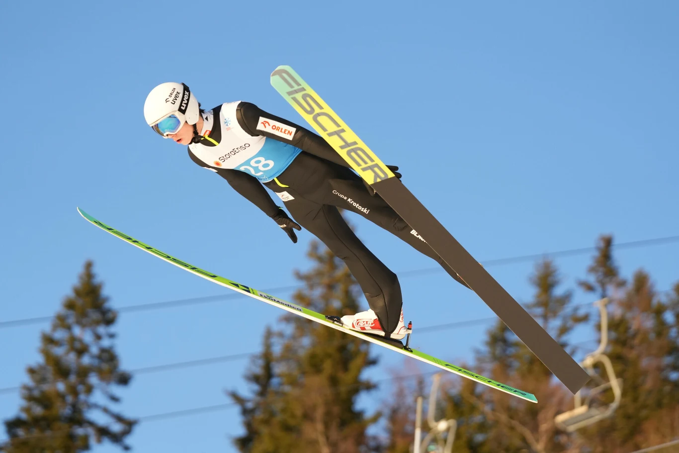 Skoczek narciarski w locie podczas zawodów, w charakterystycznej pozycji z nartami równolegle do siebie na tle nieba i rozmytych drzew w oddali.