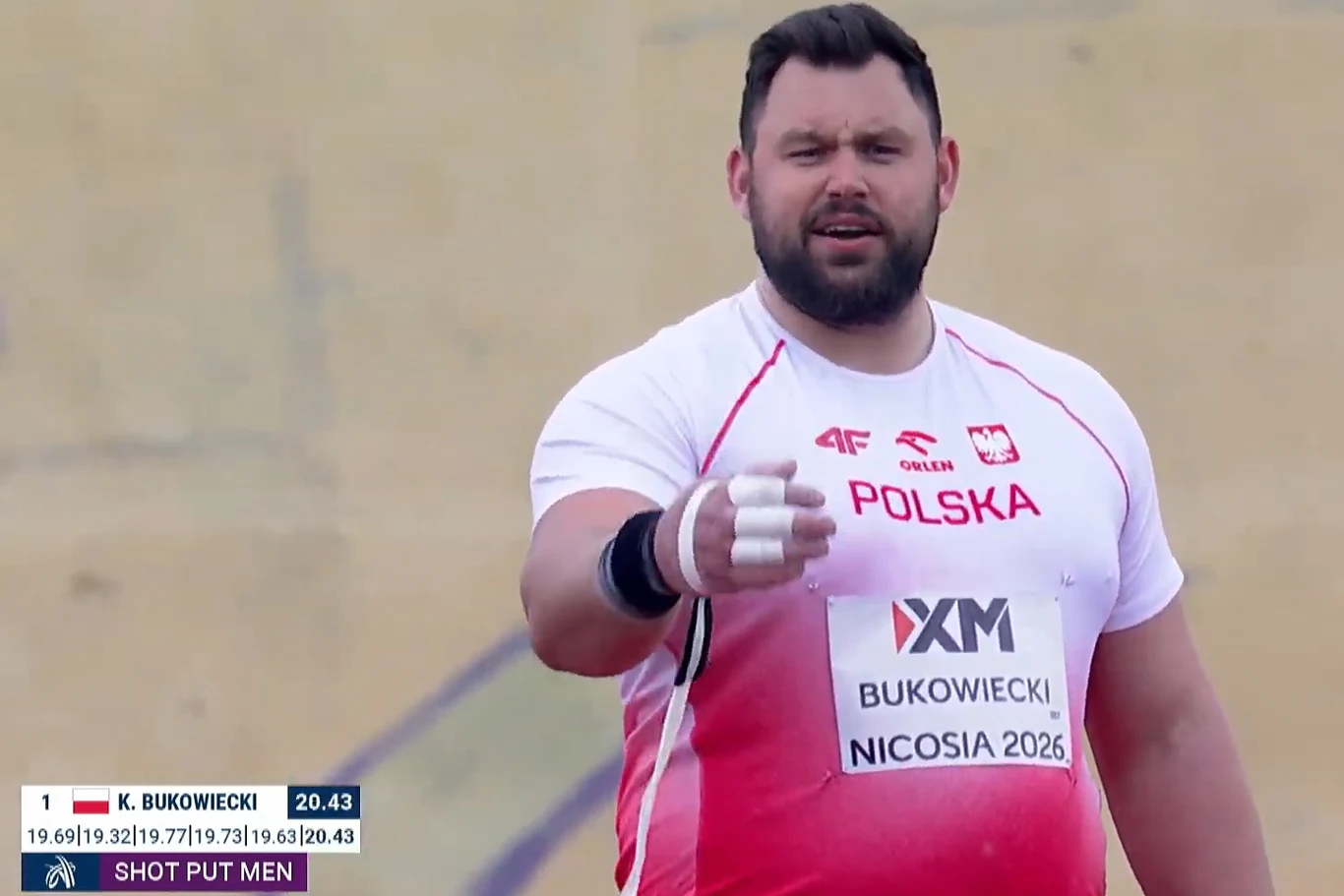 Atleta ubrany w biało-czerwoną koszulkę z napisem 'POLSKA' i godłem, przygotowany do pchnięcia kulą, skoncentrowany patrzy w stronę kamery, unosząc zaciśniętą dłoń owiniętą białą taśmą; w tle rozmyte pole stadionu lekkoatletycznego.