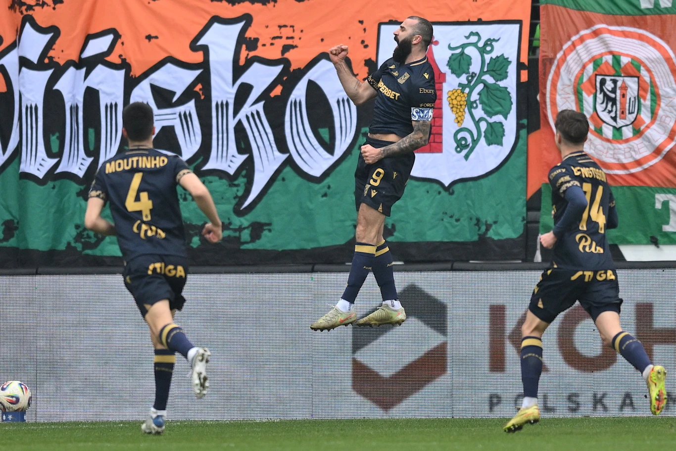 Piłkarz w granatowym stroju z numerem 9 unosi się w powietrzu celebrując bramkę, dwóch jego kolegów z drużyny biegnie w jego stronę na tle kolorowych kibicowskich flag na stadionie.