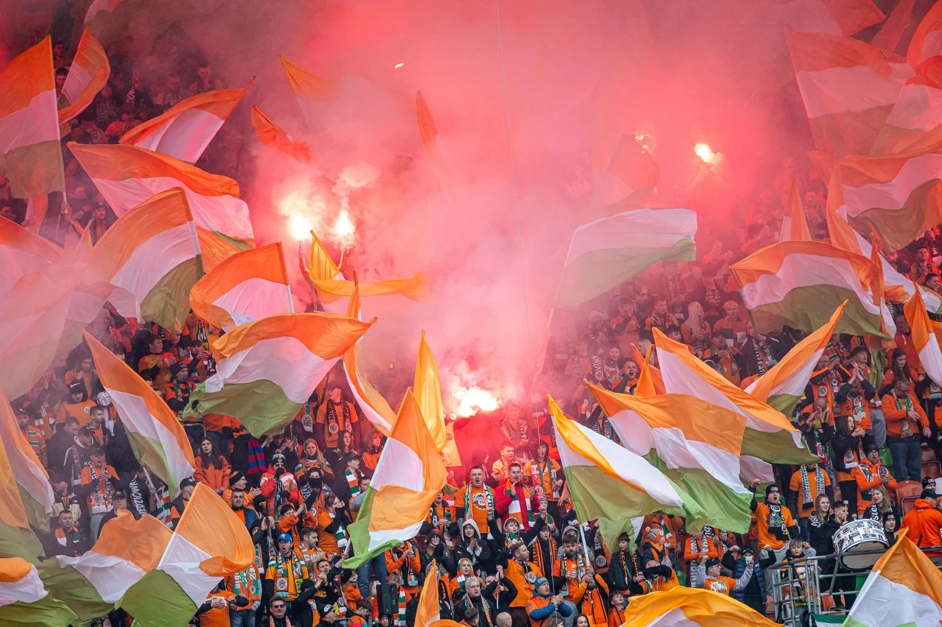 Tłum kibiców z pomarańczowo-biało-zielonymi flagami oraz odpalonymi racami tworzącymi intensywną, czerwoną poświatę nad trybuną stadionu.