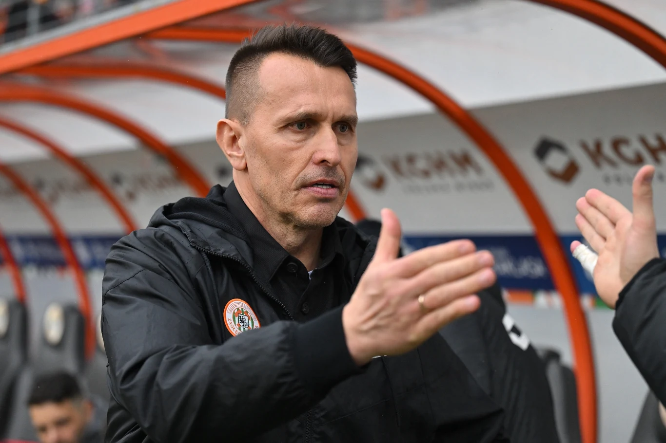 Trener w czarnej kurtce wykonuje gest powitania lub podziękowania, stojąc na tle pomarańczowej ławki rezerwowych na stadionie piłkarskim.