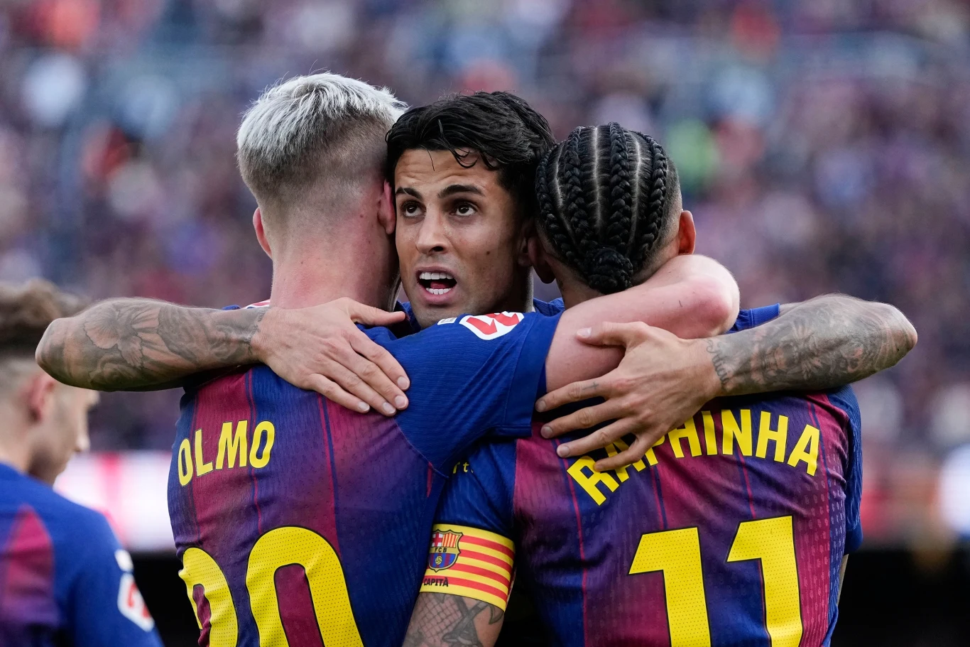 Trzech piłkarzy w koszulkach Barcelony obejmuje się po zdobyciu bramki; na pierwszym planie widoczne są nazwiska Olmo i Raphinha oraz opaska kapitańska na ramieniu środkowego zawodnika.