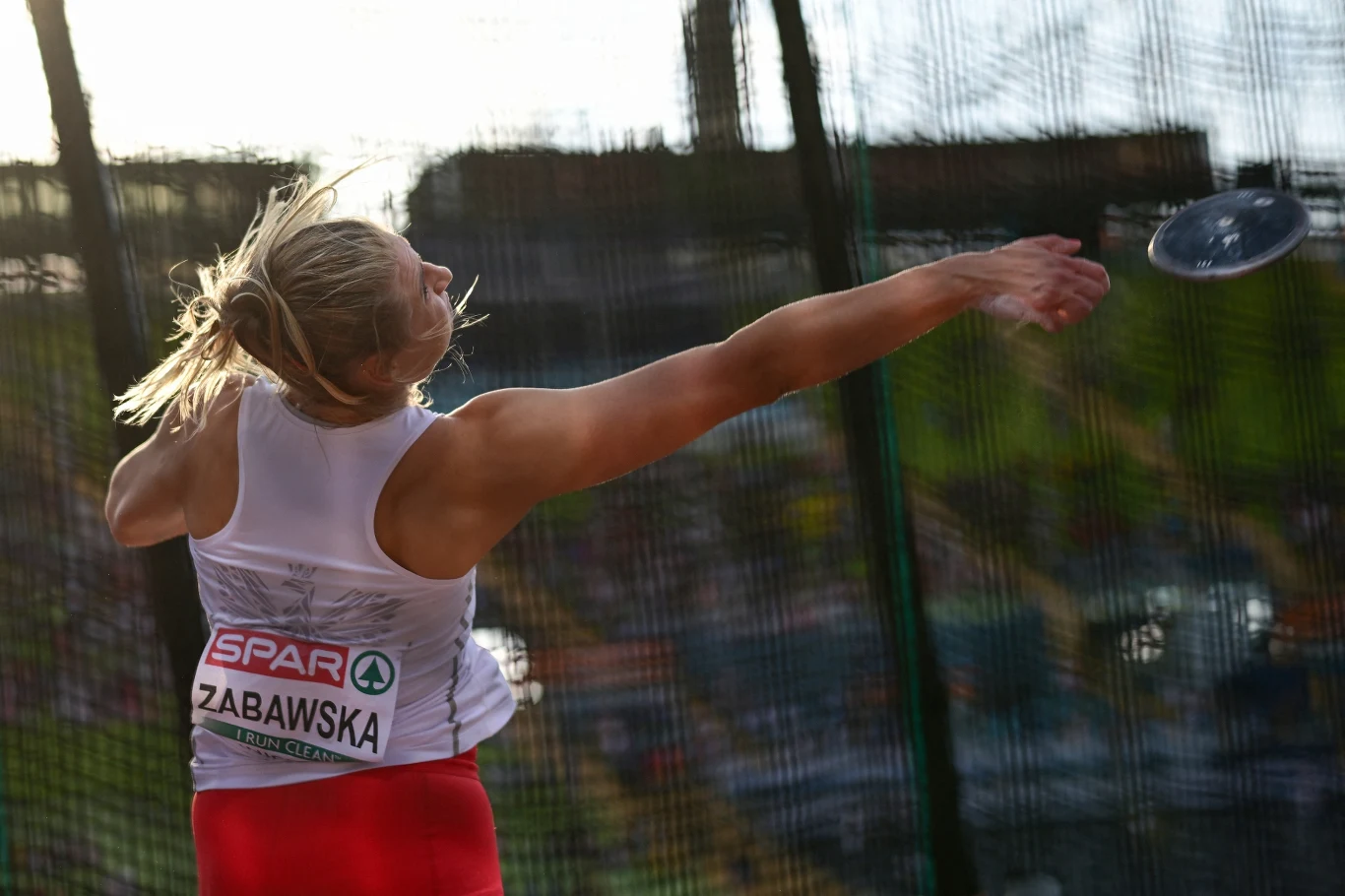 Zawodniczka w trakcie rzutu dyskiem na zawodach lekkoatletycznych, dynamiczny moment wypuszczenia dysku z rąk, sportowy strój z widocznym numerem startowym i nazwiskiem, siatka zabezpieczająca wokół klatki do rzutu.