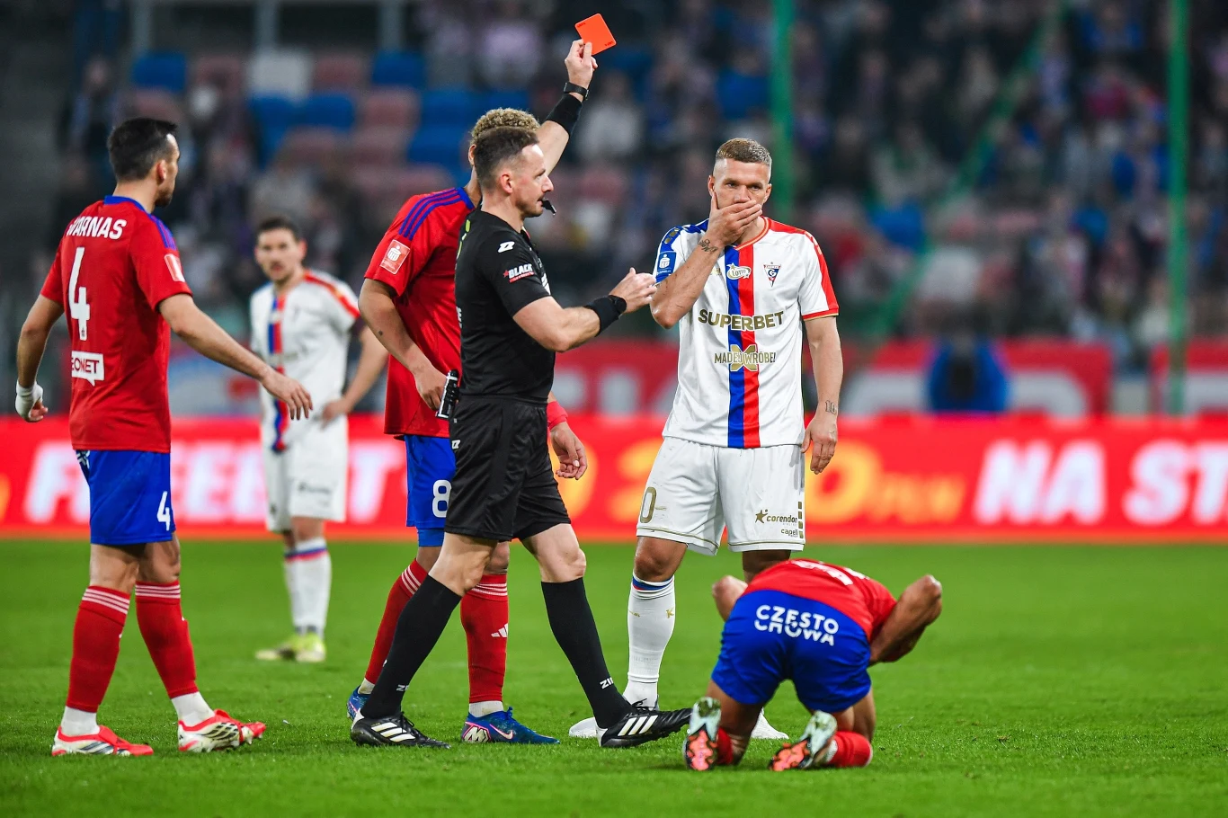 Sędzia pokazuje czerwoną kartkę piłkarzowi w białym stroju, który trzyma się za twarz, podczas gdy zawodnik drużyny przeciwnej klęczy na murawie trzymając się za nogę; wokół nich stoją inni piłkarze obserwujący sytuację.