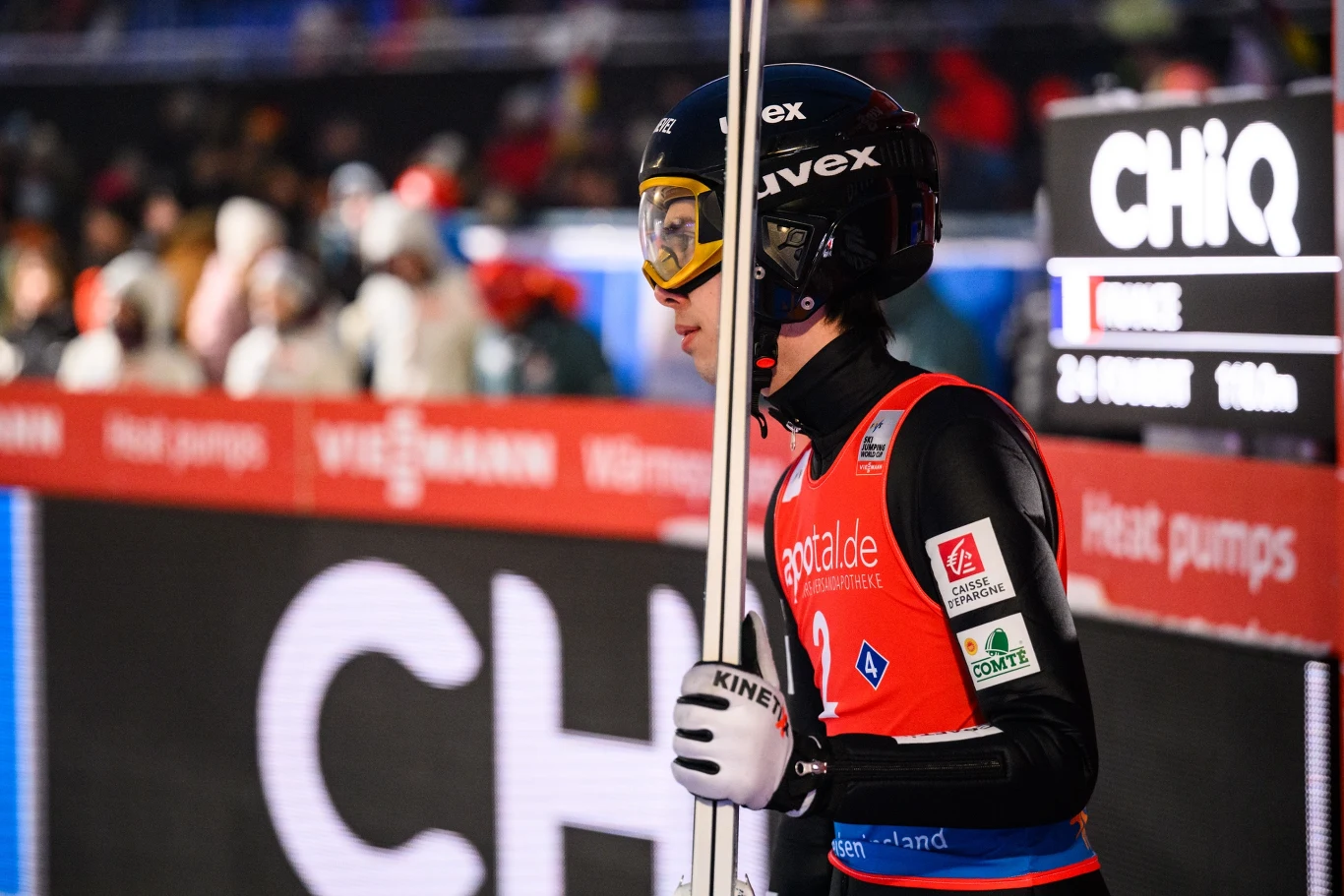 Skoczek narciarski w kombinezonie startowym i kasku z goglami trzyma narty, stojąc w strefie startowej podczas zawodów zimowych; w tle widoczna jest tablica wyników oraz zamazana publiczność.