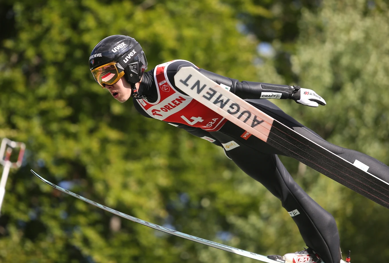 Skoczek narciarski w trakcie lotu, w aerodynamicznej pozycji z wyciągniętymi ramionami, ubrany w czarny kombinezon z numerem startowym i kaskiem, na tle zielonych drzew.