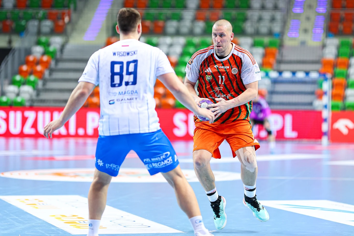 Zagłębie – Stal Dwaj zawodnicy piłki ręcznej w akcji na hali sportowej, gracz w pomarańczowym stroju prowadzi piłkę, podczas gdy przeciwnik w białoniebieskim zestawie próbuje go zatrzymać.