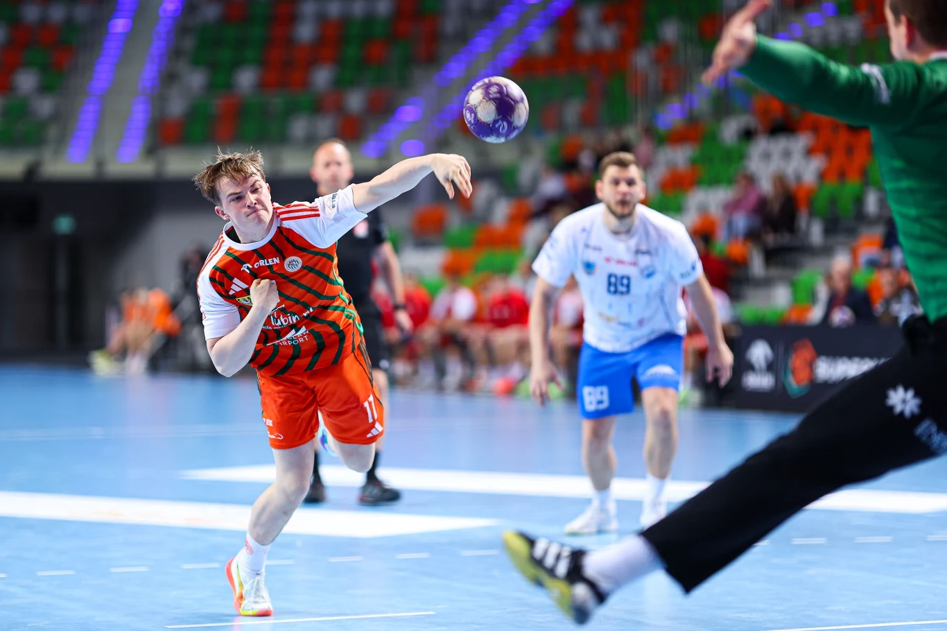 Zagłębie – Stal Zawodnik w pomarańczowym stroju rzuca piłkę podczas meczu piłki ręcznej, naprzeciwko bramkarz w zielonym ubraniu przygotowuje się do obrony; w tle widoczni są inni zawodnicy i trybuny z kolorowymi siedziskami.