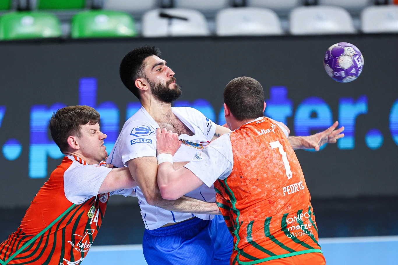 Zagłębie – Stal Dwóch zawodników drużyny w pomarańczowych koszulkach próbuje powstrzymać zawodnika drużyny przeciwnej ubranego na biało-niebiesko, podczas dynamicznej akcji z piłką ręczną na hali sportowej.