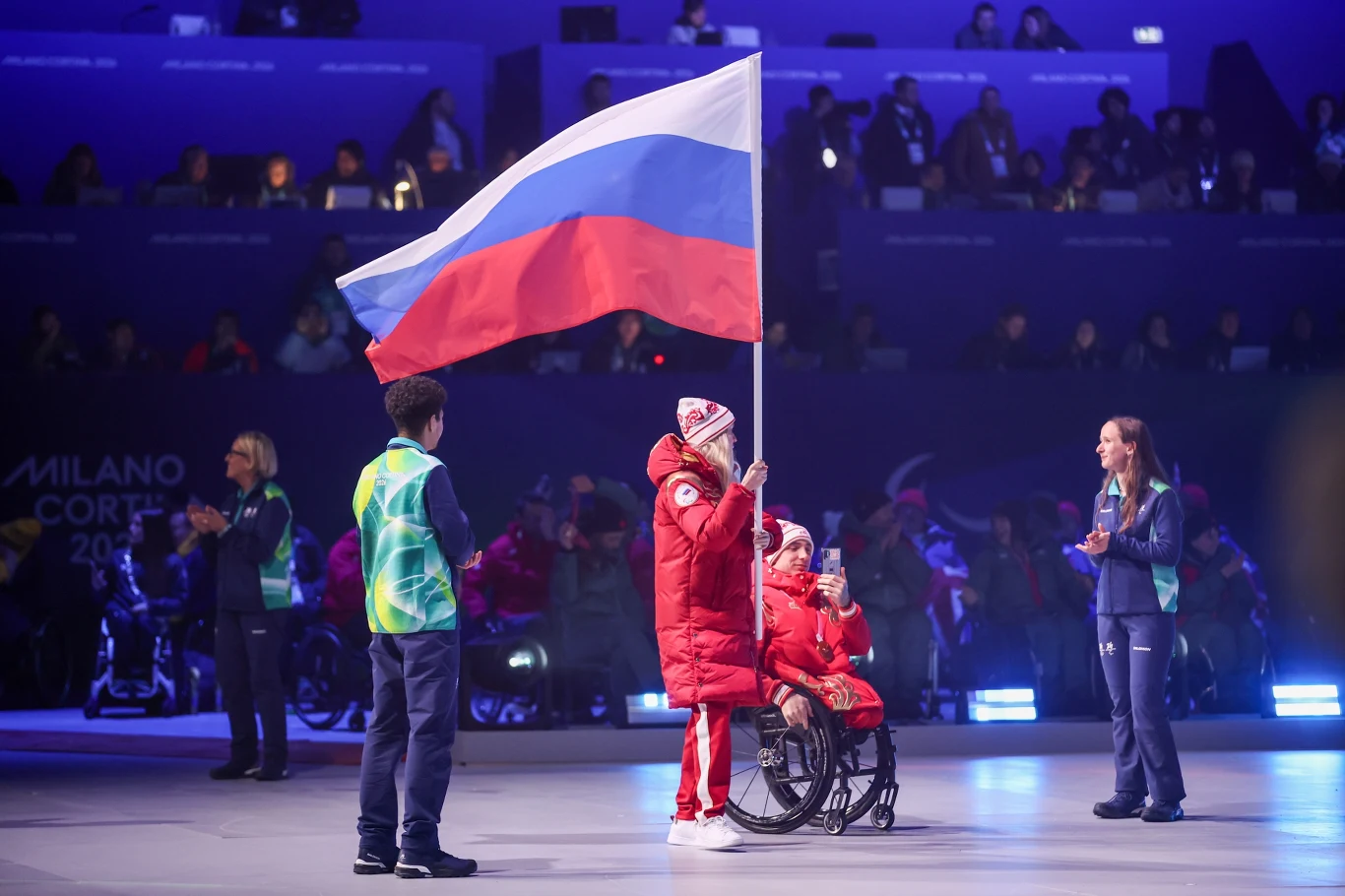 Sportowiec na wózku inwalidzkim ubrany w czerwony strój zimowy trzyma dużą rosyjską flagę, obok stoją inni uczestnicy w sportowych strojach, a w tle widoczna scena z napisem Milano Cortina.
