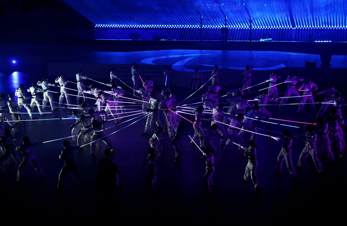 Grupa tancerzy w białych strojach wykonuje synchroniczny układ ze świetlnymi, kolorowymi kijami, tworząc efektowne wzory na ciemnej scenie oświetlonej błękitnym światłem.