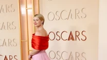 Kobieta w eleganckiej sukni z długim, różowym trenem i czerwonym gorsetem pozuje na czerwonym dywanie przed napisem 'Oscars', z subtelnym uśmiechem i upiętymi blond włosami.