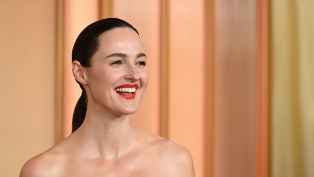 Uśmiechnięta kobieta w czerwonej sukience z odkrytymi ramionami, o ciemnych, gładko zaczesanych do tyłu włosach, stoi na tle w ciepłych kolorach.