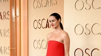 Kobieta w asymetrycznej, czerwonej sukni z długim trenem i wysokim rozcięciem, stojąca na czerwonym dywanie, w tle ściana z napisem 'OSCARS'.
