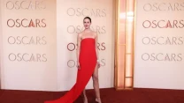 Kobieta w efektownej czerwonej sukni z długim trenem, stojąca na czerwonym dywanie ceremonii Oscarów, na tle ściany z powtarzającym się napisem 'Oscars'.