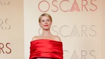 Kobieta w eleganckiej, długiej sukni z różowym dołem i czerwonym, szerokim marszczonym topem, stoi na czerwonym dywanie na tle ściany z napisem OSCARS.