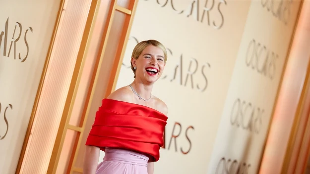 Uśmiechnięta kobieta w eleganckiej sukni z czerwonym gorsetem i różową spódnicą pozuje na tle ściany z napisem OSCARS.