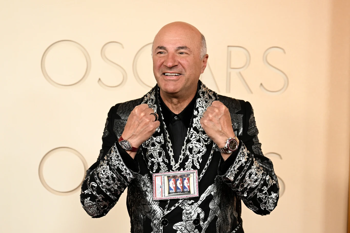 Uśmiechnięty mężczyzna ubrany w czarną marynarkę z bogatym srebrnym wzorem i wieloma srebrnymi naszyjnikami, na jednym z nich zawieszona ramka z kartami do gry, na tle napisu 'OSCARS'.