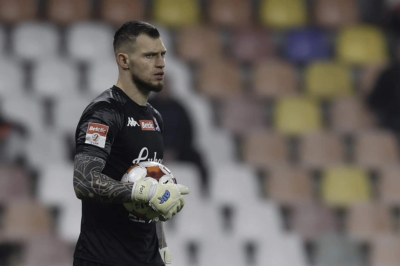 Bramkarz w czarnym stroju z piłką w rękach, stojący na tle pustych, wielokolorowych trybun stadionu.