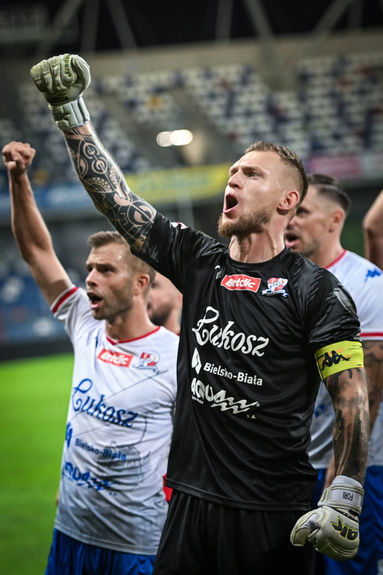 Grupa piłkarzy w strojach meczowych świętuje zwycięstwo na stadionie, na pierwszym planie bramkarz z opaską kapitańską i uniesioną zaciśniętą pięścią, wyrażający silne emocje i triumf.