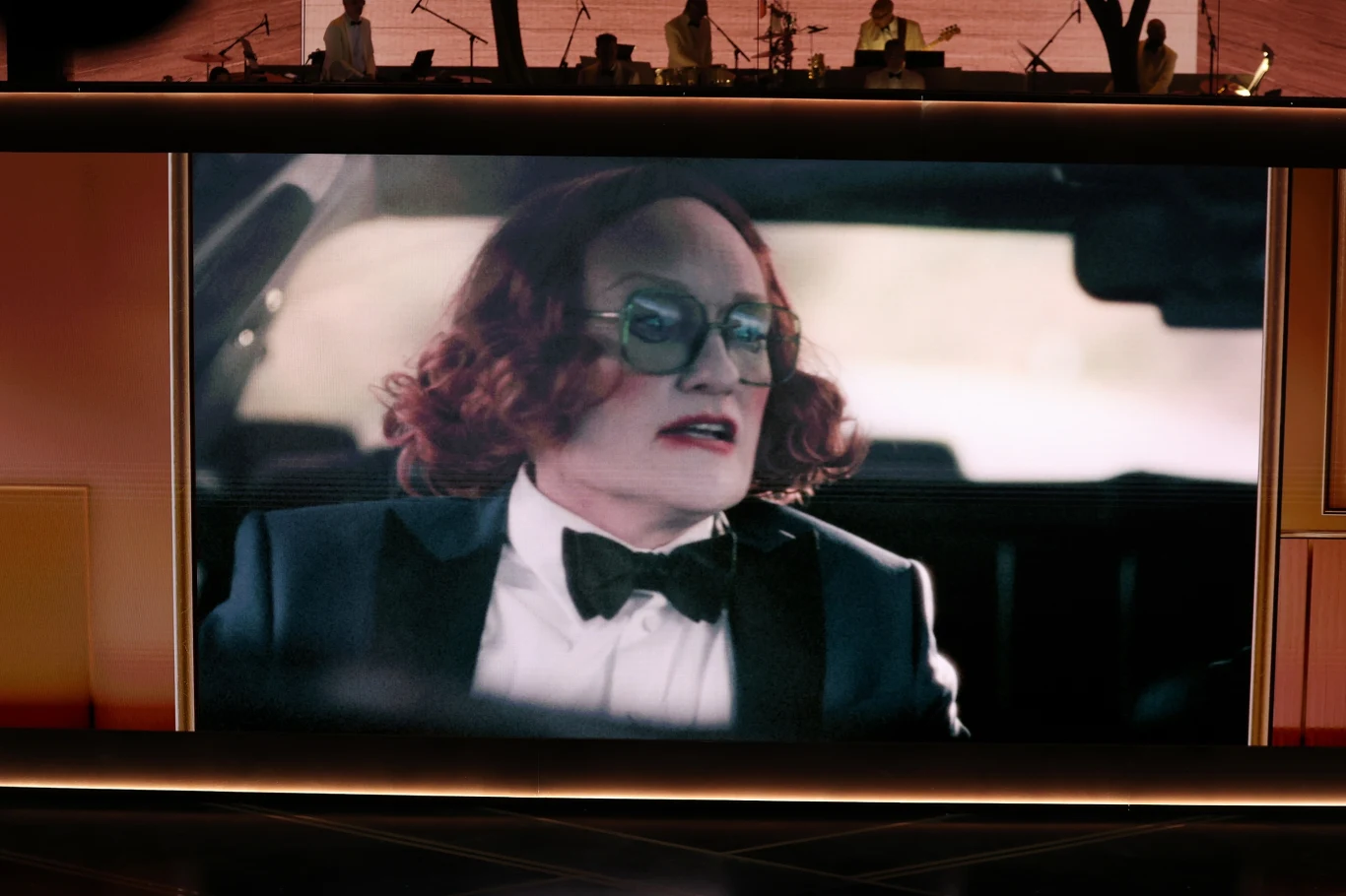Osoba w peruce o rudych, kręconych włosach i eleganckim smokingu z muchą siedzi za kierownicą samochodu, patrząc w bok i nosząc duże okulary przeciwsłoneczne; obraz wyświetlany jest na dużym ekranie, na górze widoczne są sylwetki muzyków na scenie.